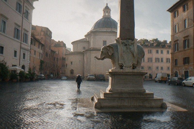 Morning drizzle on the granite cobblestones of Piazza della Minerva in Rome, Italy, Bernini's elephant obelisk rising wet and dark in the frame, one distant figure with their back turned crossing toward the pantheon-like dome, puddles reflecting grey light.