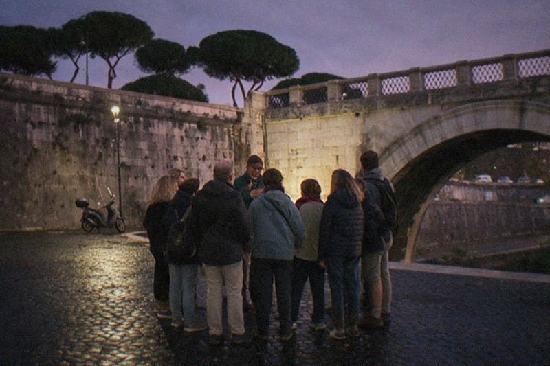 A small tour group huddles close around their guide on the Tiber embankment in Rome, Italy, at dusk beneath the stone arches of Ponte Sant'Angelo, umbrella pines dark against a violet sky, wet cobblestones reflecting a lone streetlamp.