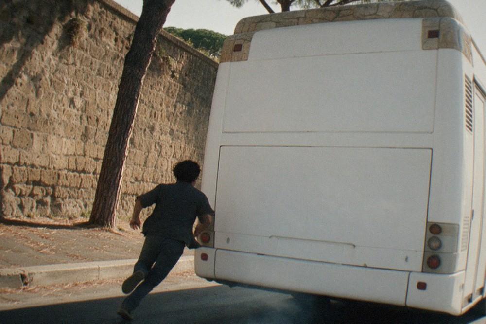 A Rome bus pulling away from a stop beside a stone wall and umbrella pines