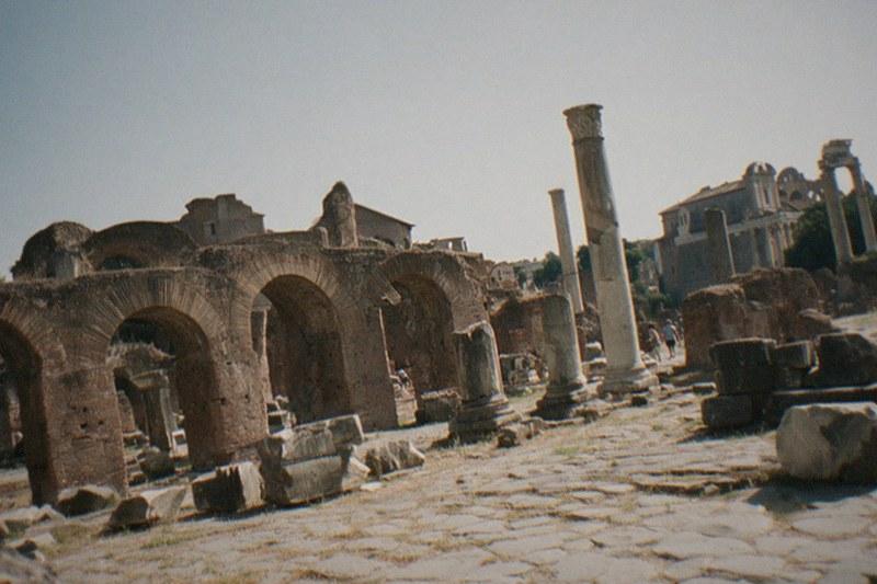 Arcos de ladrillo desgastado y columnas de mármol rotas del Foro Romano bajo un cielo brumoso de tarde, hierba seca asomando entre el pavimento agrietado.