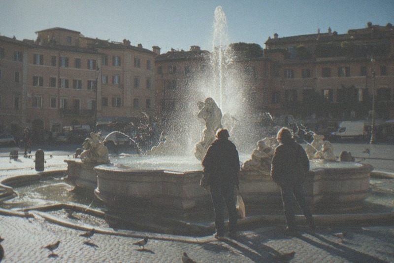 Una fuente barroca con sus tritones de piedra en la plaza Navona, el sol de media mañana ilumina la pila húmeda, palomas dispersándose sobre los sampietrini, bajo los palacios romanos de color ocre.