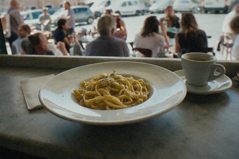 Pasta fresca y una taza pequeña de espresso sobre un mostrador de mármol gastado en un local sencillo de Roma, el comedor concurrido y la plaza exterior se desdibujan en el fondo.