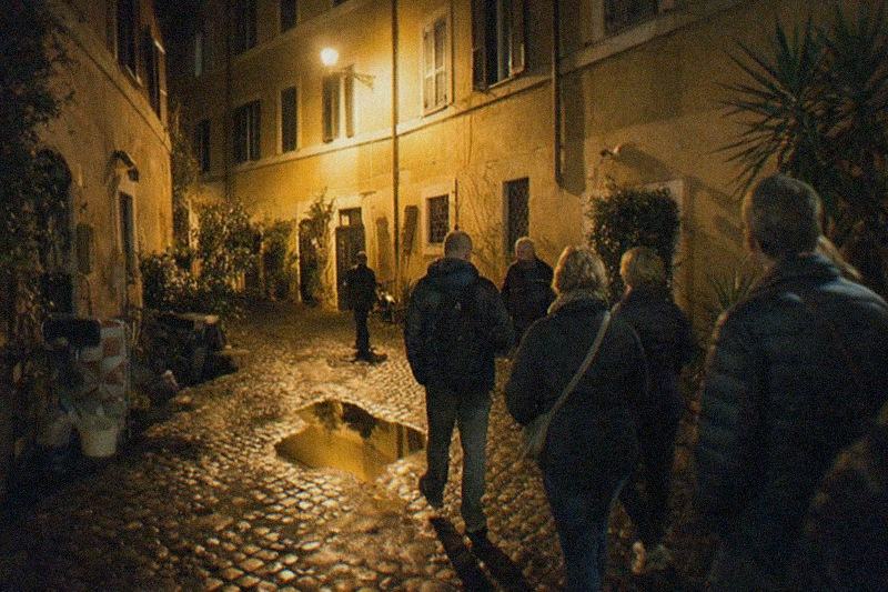 Un callejón estrecho de Trastevere de noche, los sampietrini mojados reflejan la luz ámbar de las farolas bajo paredes ocre con postigos cerrados, un charco que refleja la única farola de la calle.