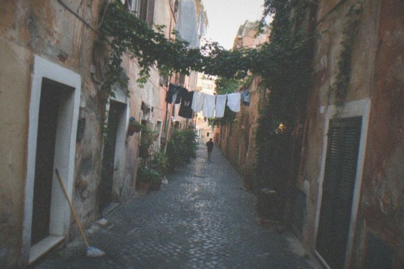 Hiedra y tendederos se cruzan sobre un callejón empedrado de Trastevere, el yeso ocre y oxidado se desprende de las paredes viejas, una figura camina alejándose por el callejón a la luz del atardecer.