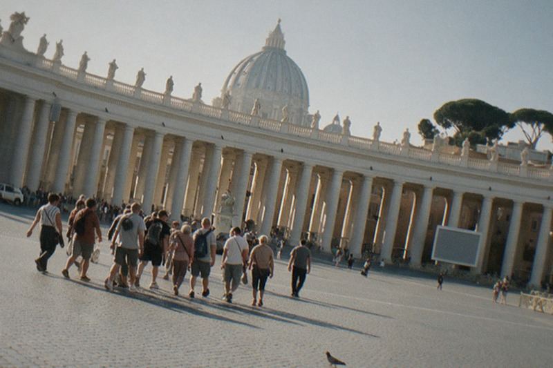 La columnata curva de Bernini y la cúpula pálida de San Pedro sobre la amplia plaza empedrada, bajo una luz plana de primera hora, con pinos piñoneros al fondo y una paloma cruzando el espacio vacío.