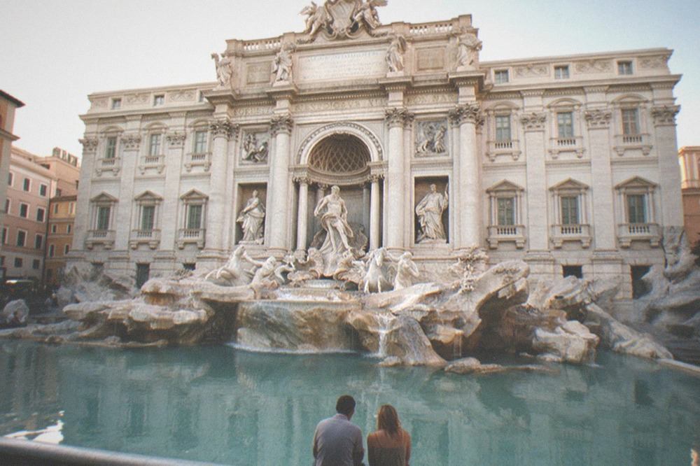 The Trevi Fountain at dusk, with the square mostly empty, the lit Baroque façade of Palazzo Poli reflecting in the blue water, and a couple seated at the basin edge.