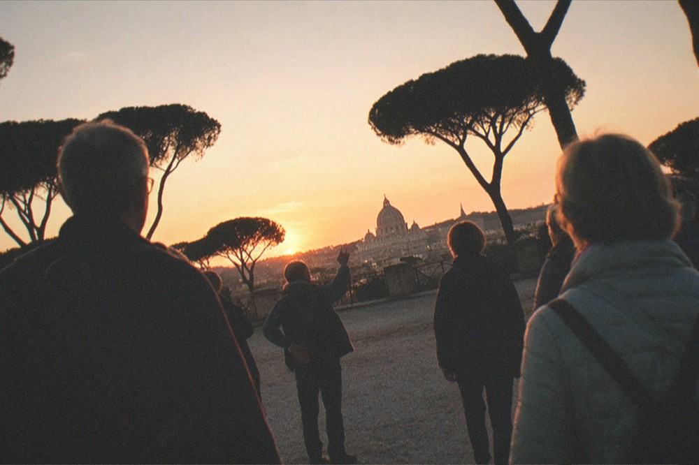A group of walkers on the Pincio terrace at sunset with a guide pointing toward the Vatican, Villa Borghese's stone pines silhouetted against the orange sky.