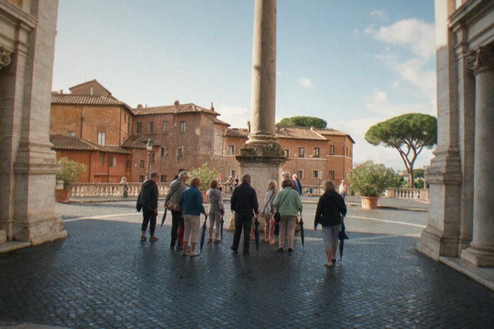 A small group of walkers gathered around a guide beside an ancient stone column in a Roman square