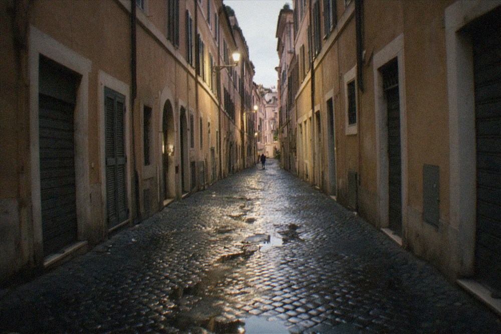 A quiet Rome street after rain, with reflections of street lamps on wet cobblestones.