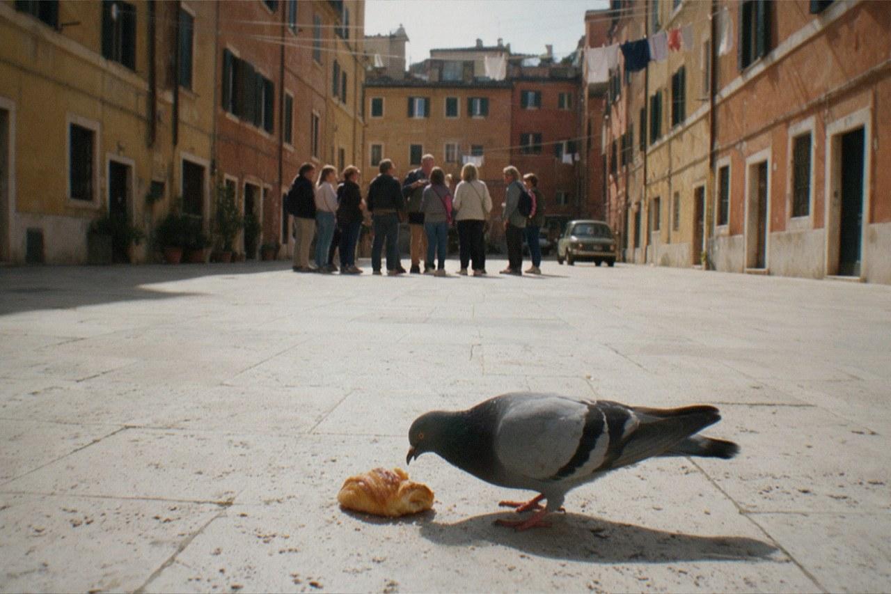 Un pequeño grupo de tour a pie se alinea al fondo de una plaza empedrada de travertino en Roma, de espaldas a su guía en una luminosa mañana de primavera, con fachadas ocre y terracota y tendederos colgantes llenando el encuadre tras ellos.